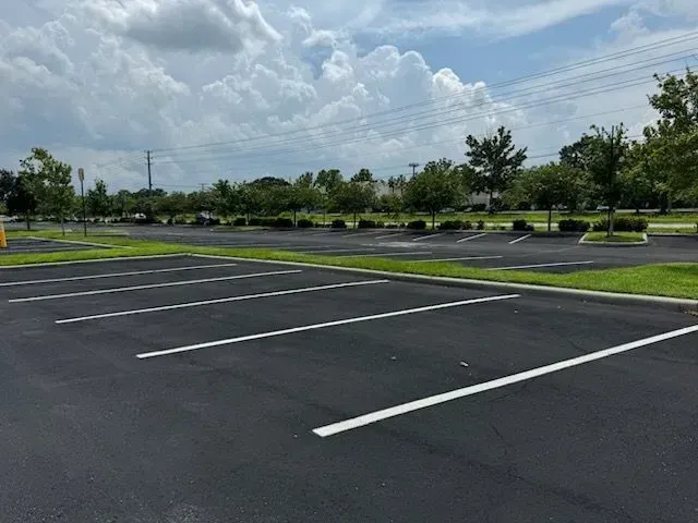 Empty parking lot with white lines under a partly cloudy sky, bordered by trees and grass