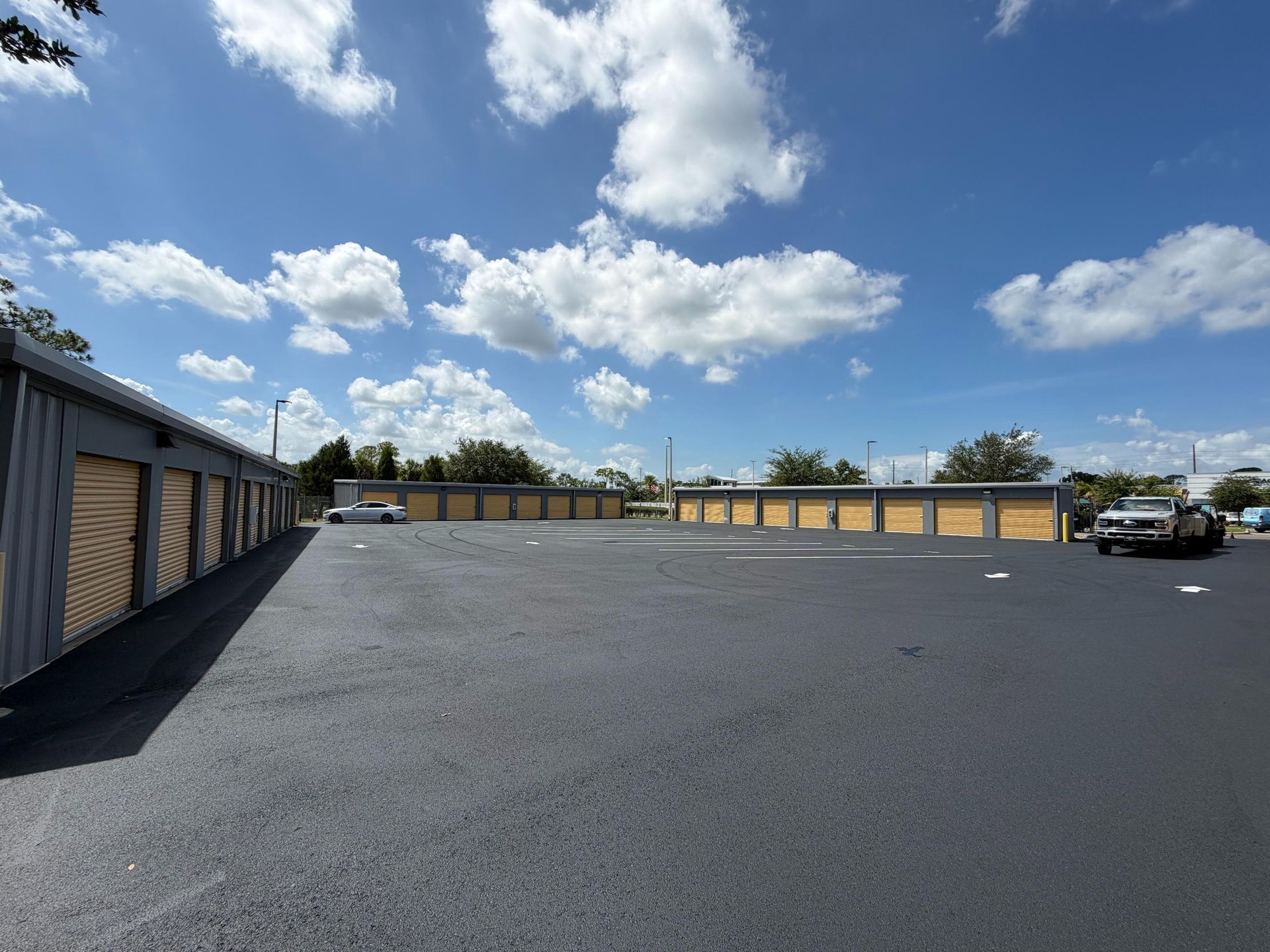 Wide gravel lot with storage units under a bright blue sky with scattered clouds.