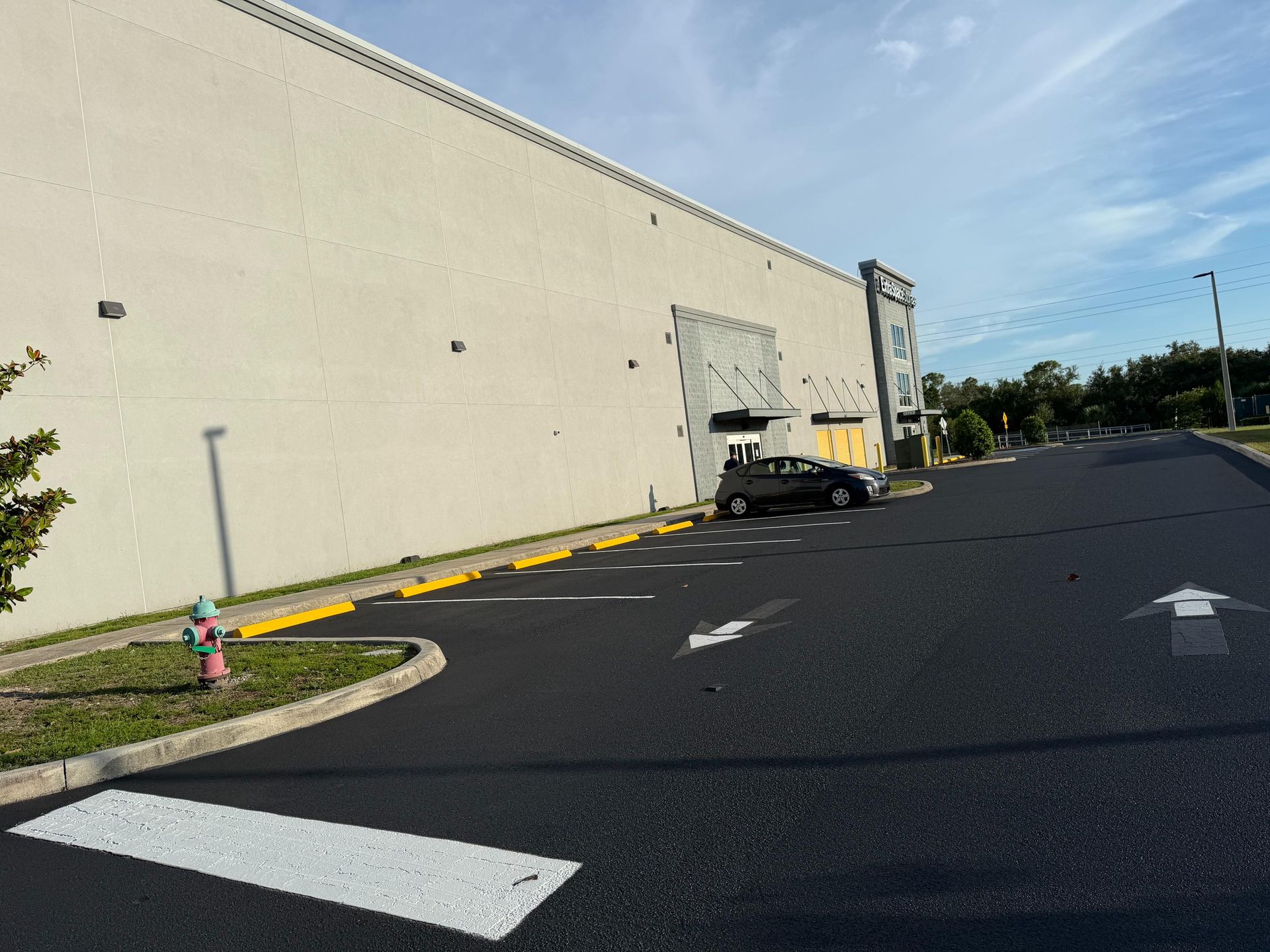 Empty parking lot beside a beige warehouse, with a truck at a loading dock and arrows painted on the asphalt.