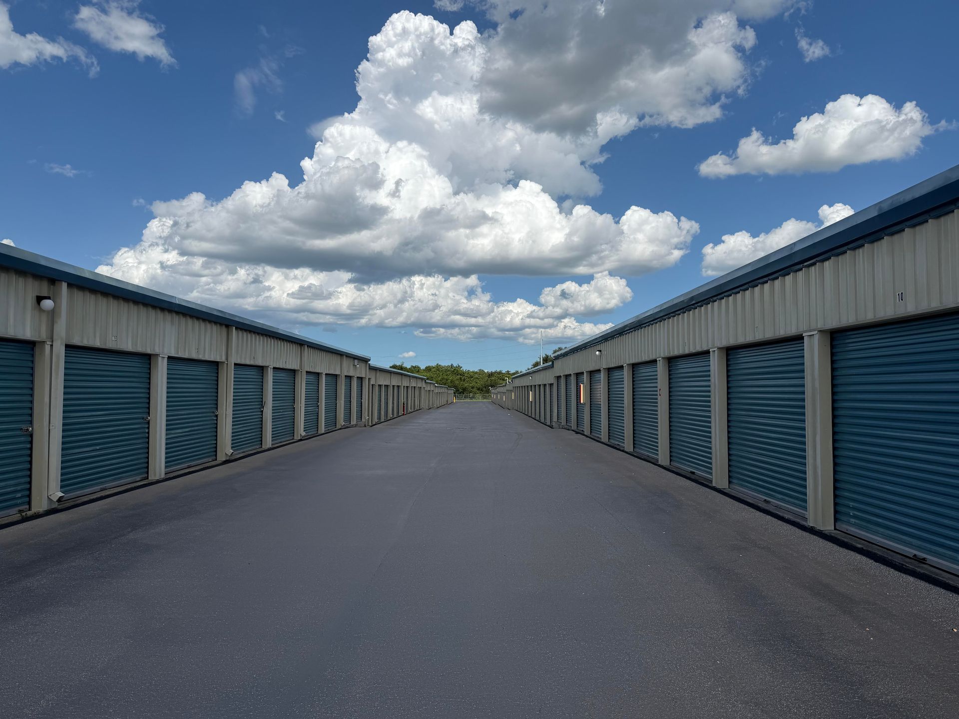 Wide paved lane between rows of blue storage units under a bright cloudy sky