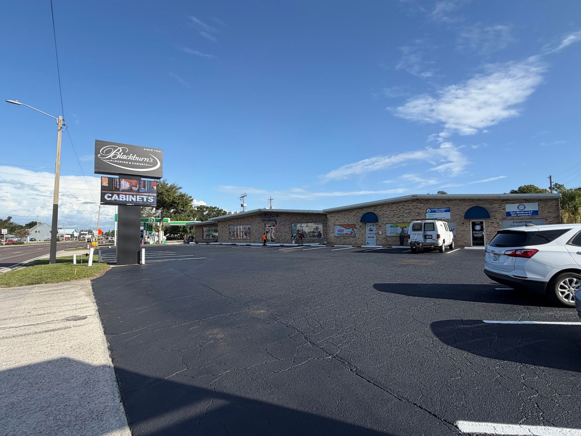 Parking lot beside a roadside building under a blue sky, with cars parked and a sign by the entrance.