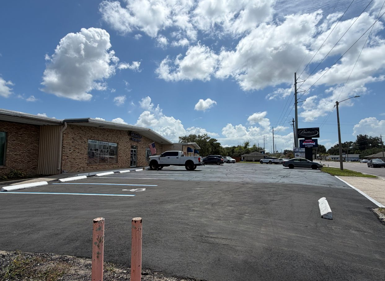 Parking lot beside a low brick building under a bright blue sky with clouds, with a white pickup truck and signposts.