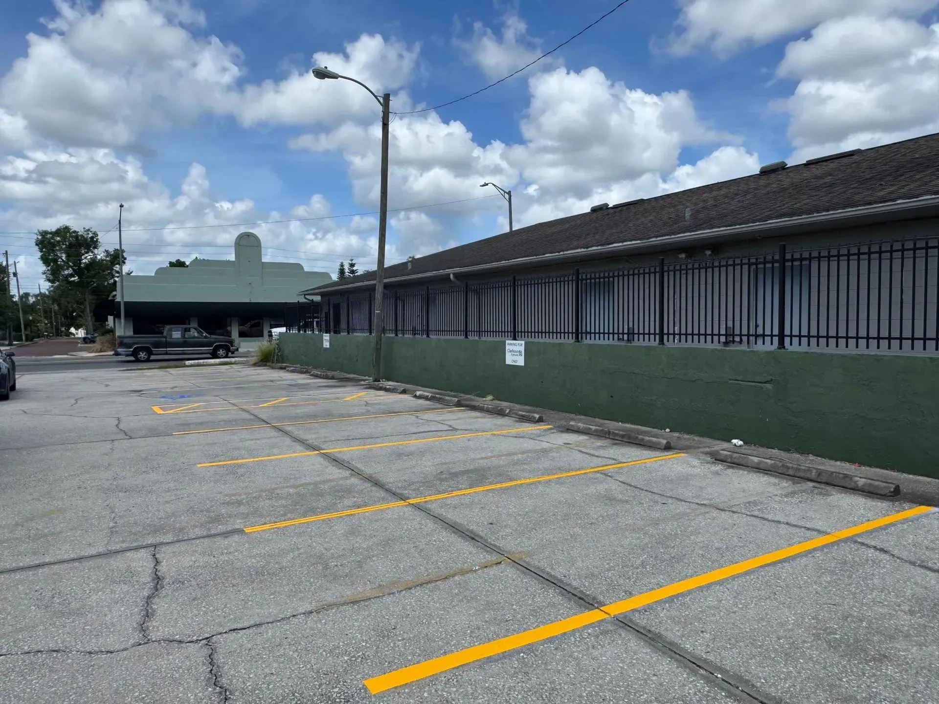 Empty parking lot beside a long building with a green fence under a cloudy sky