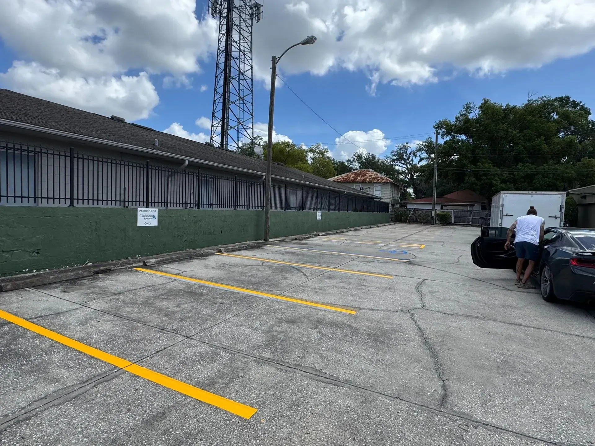 Empty parking lot with yellow lines, a person beside a parked car, and a green fence under a cloudy sky