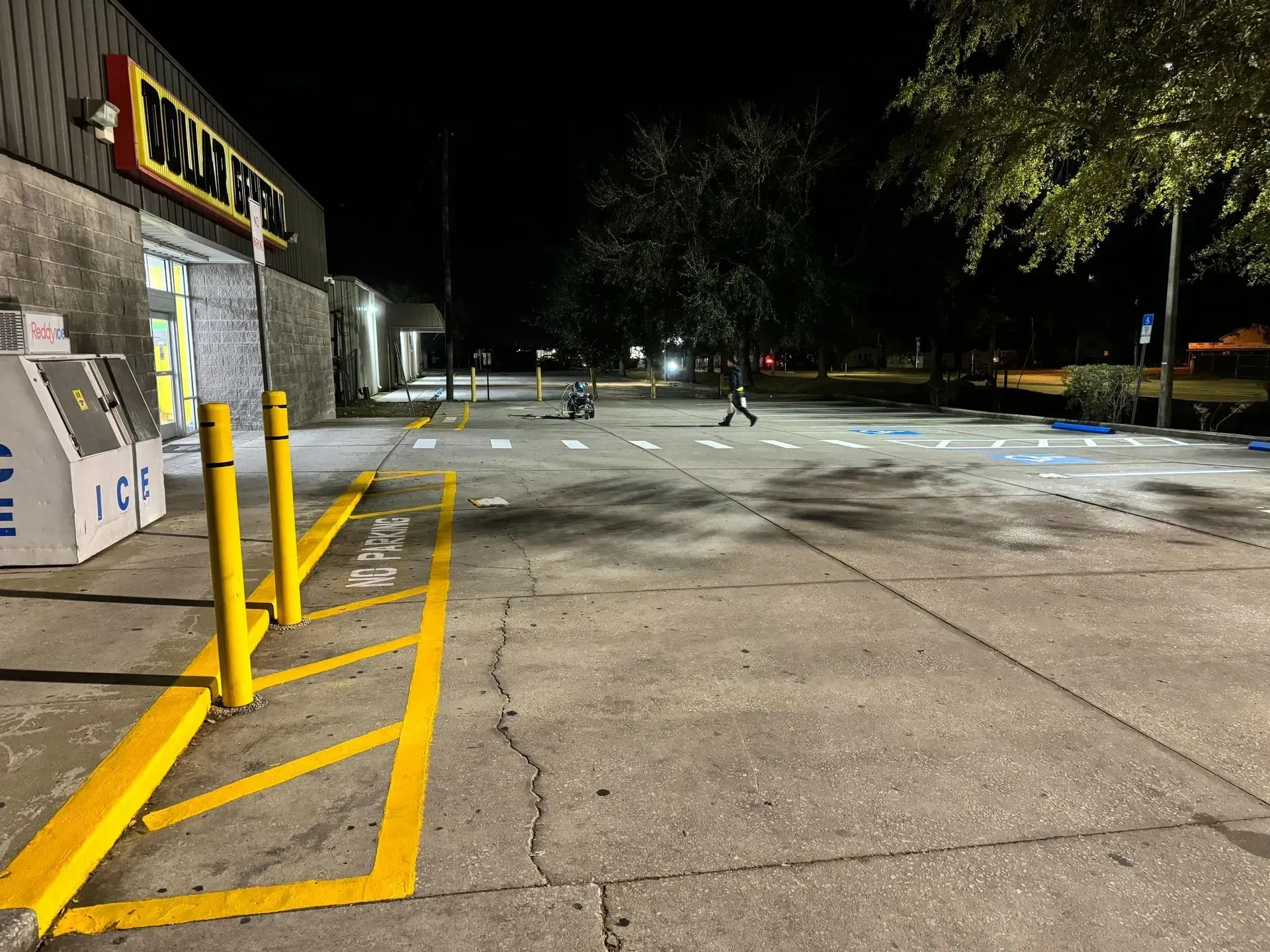 Nighttime parking lot with a store on the left, yellow barriers, and a few people in the distance.