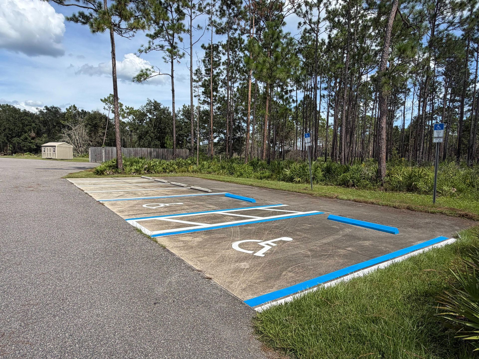 Empty accessible parking space with blue lines and wheelchair symbol beside a paved lot and trees