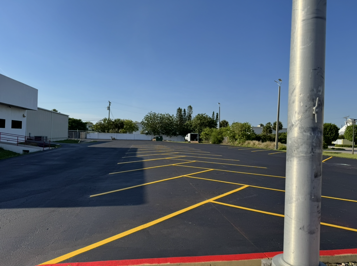 Empty parking lot with yellow lines, a white building on the left, and a metal pole in the foreground.