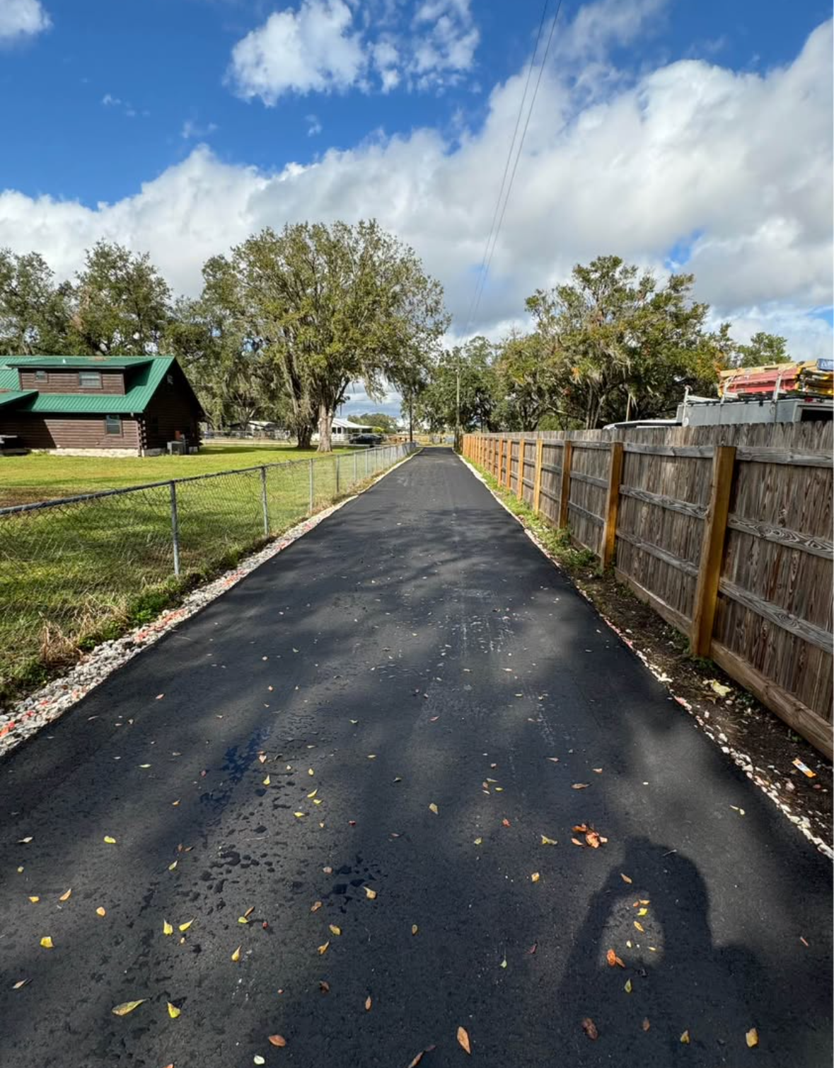 New black asphalt path between a wooden fence and grassy yard under a partly cloudy sky
