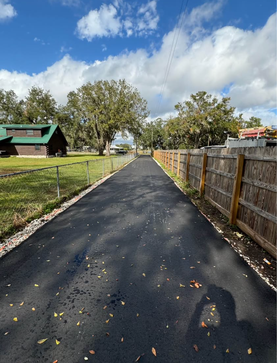 Paved driveway beside wooden fence and green lawn, leading toward trees under a partly cloudy sky