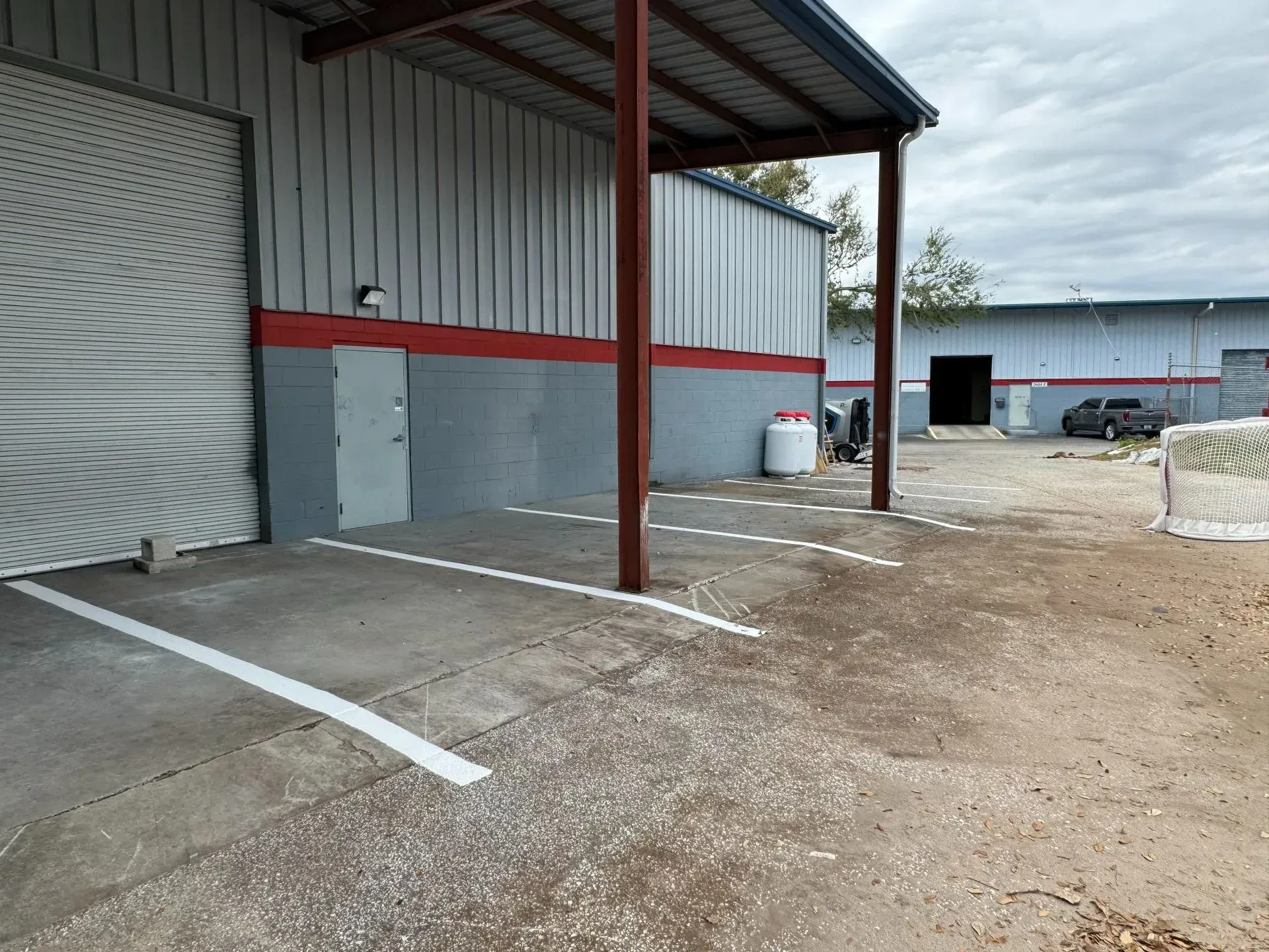 Empty industrial loading area with metal building, red beams, and gravel parking spaces under cloudy sky