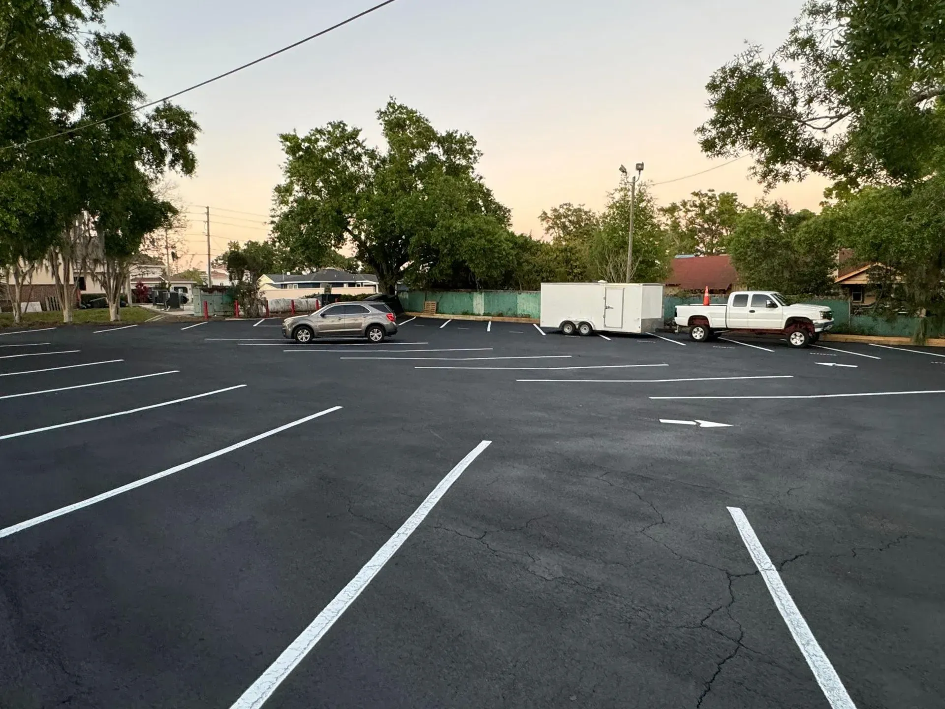 Empty parking lot at dusk with white lane markings, parked cars, and trees along the edge.