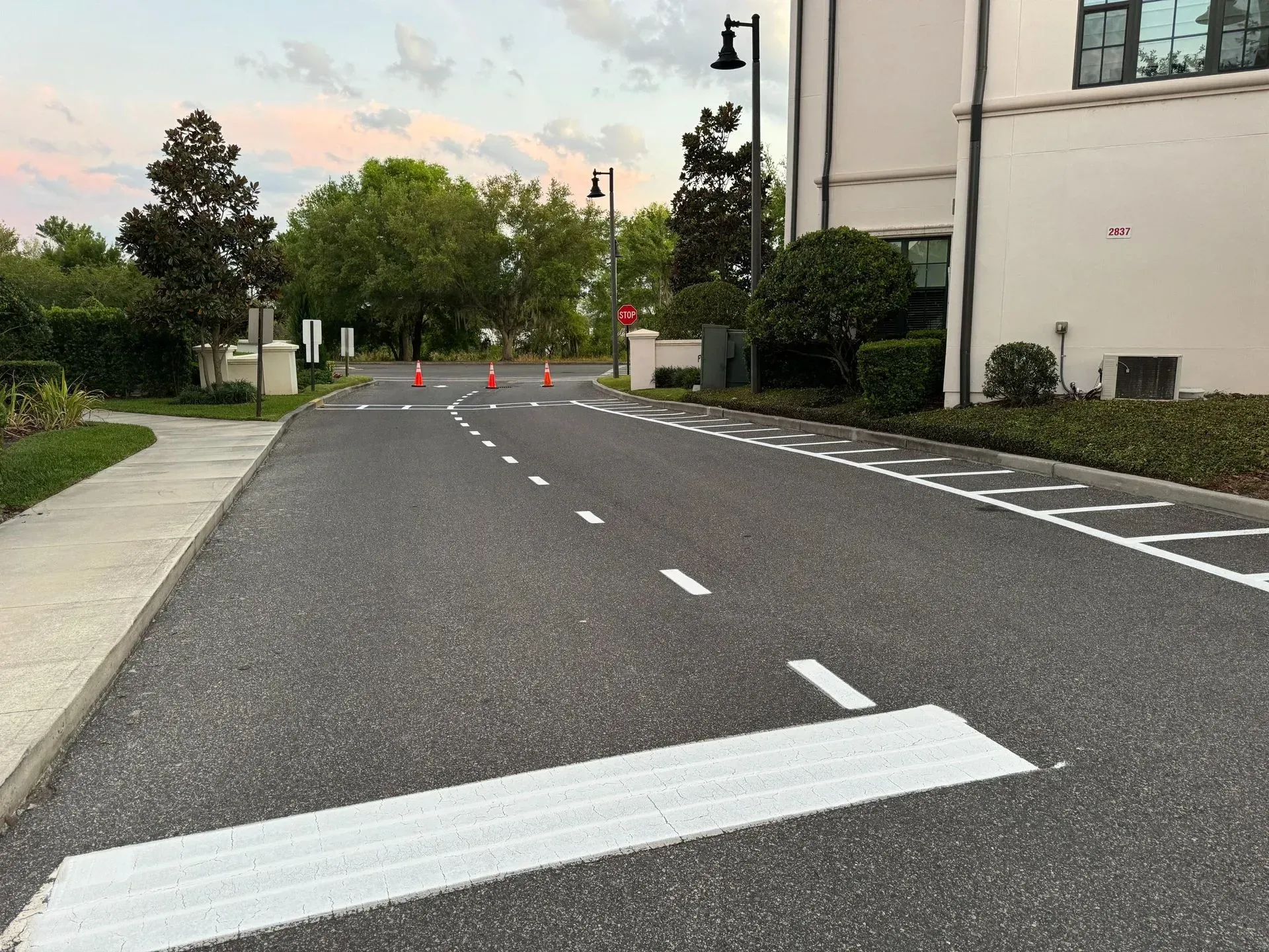 Empty paved road with white lane markings, orange cones, and buildings along the sides at dusk