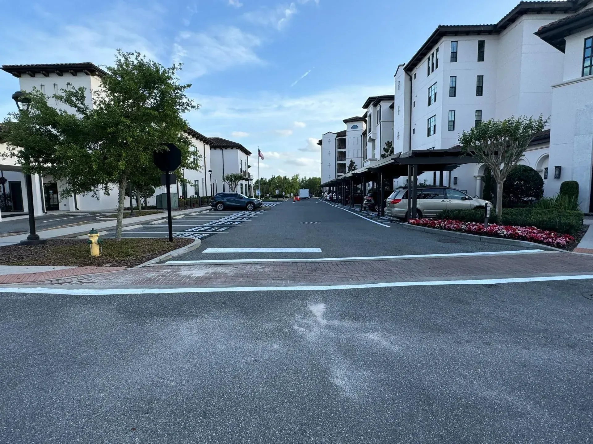 Residential street with white apartment buildings, trees, and flower beds under a blue sky