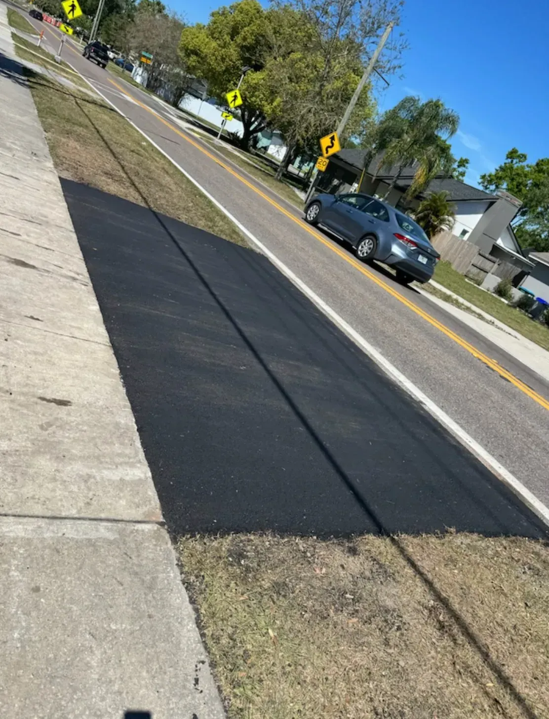 Sidewalk beside a road with a fresh black asphalt strip and a car driving past houses and trees.