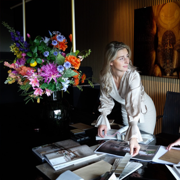 Een vrouw schikt tijdschriftpagina's naast een groot, kleurrijk bloemstuk in een schemerig verlichte kamer.