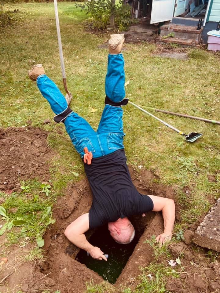 A person wearing a black shirt and blue pants is upside down, head and arms inside a rectangular hole in the ground.