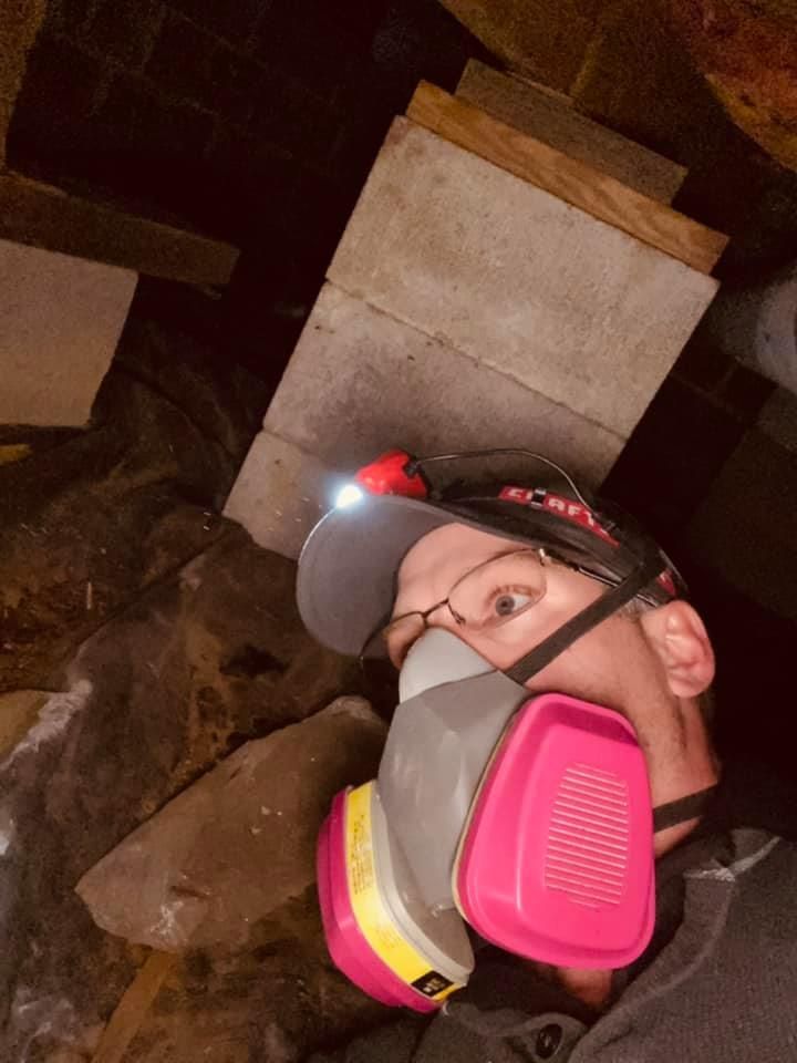 A person wearing a respirator mask and headlamp in a dark attic, looking toward a cinder block support structure.