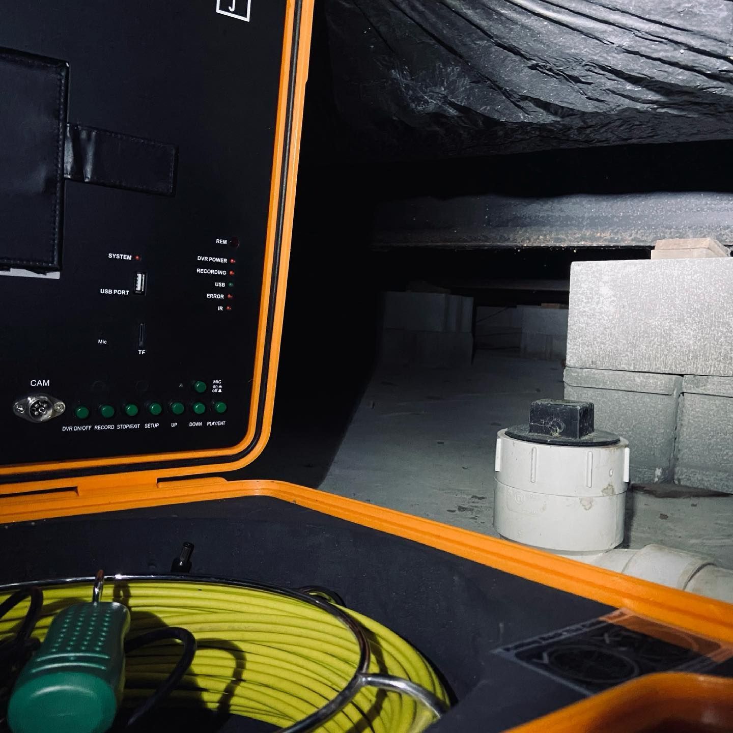 A plumbing inspection camera case sits open in a dark crawlspace next to a white PVC cleanout pipe.