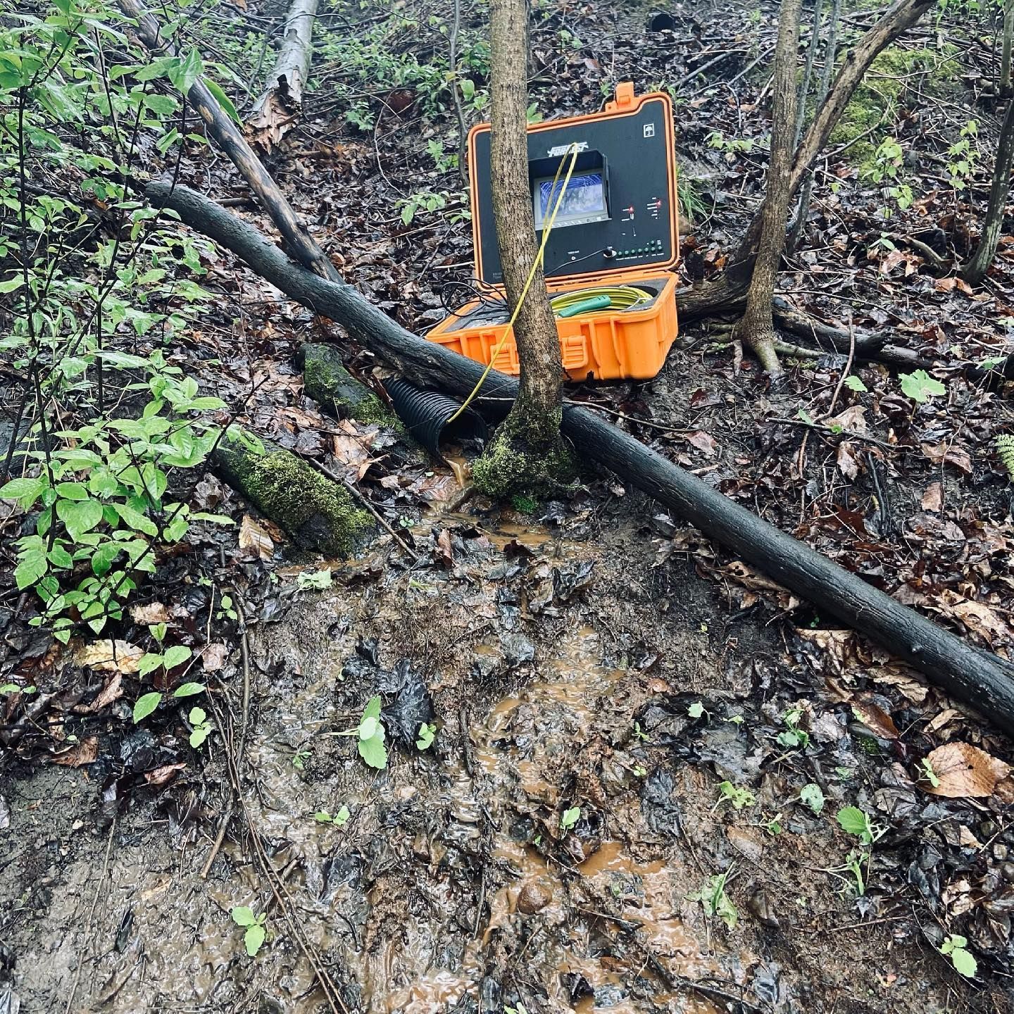 An orange portable diagnostic monitor is positioned on wet, muddy ground between trees near a black pipe in a wooded area.