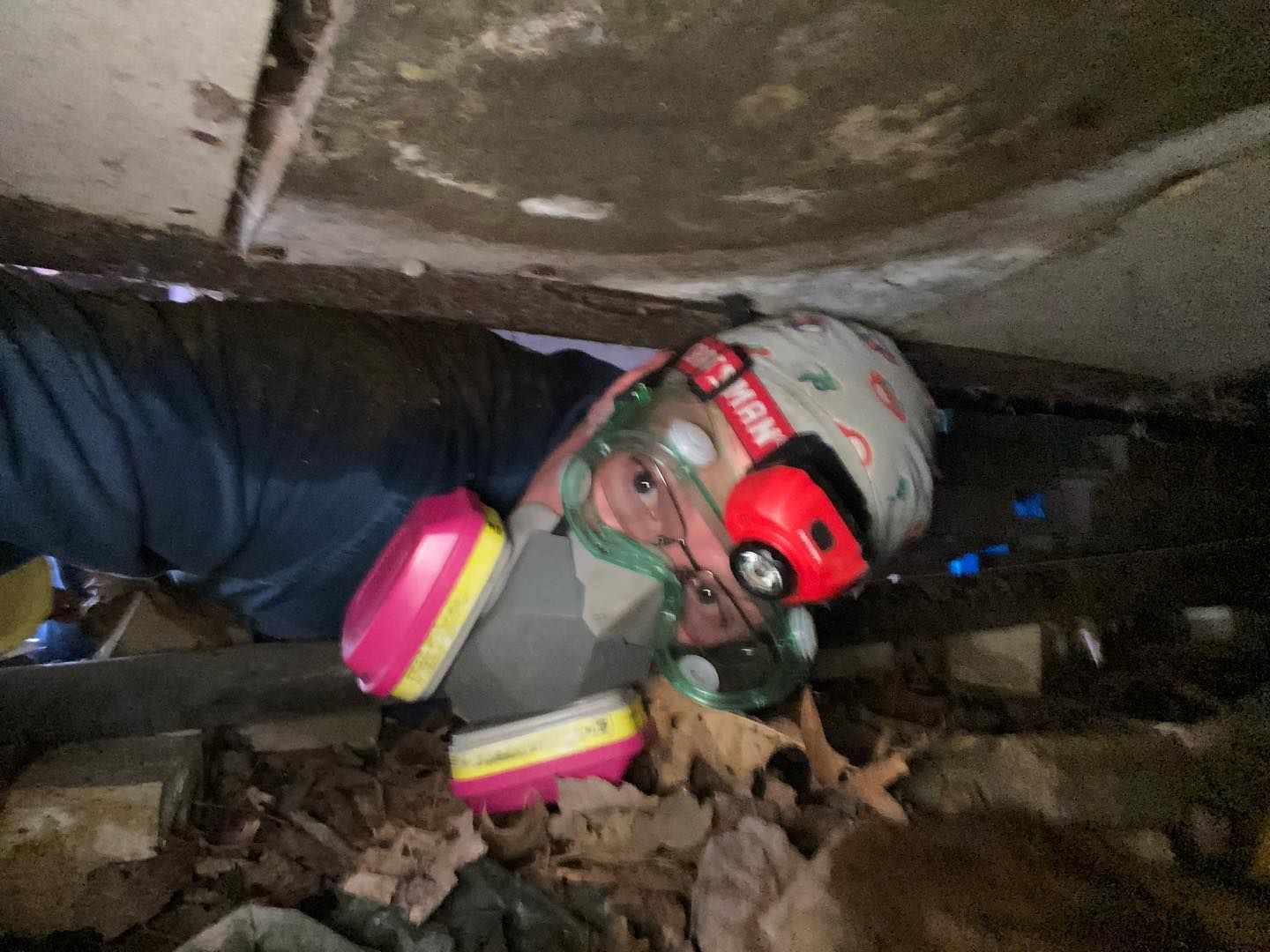 A person wearing a respirator and headlamp crawls through a tight, debris-filled crawl space beneath a structure.