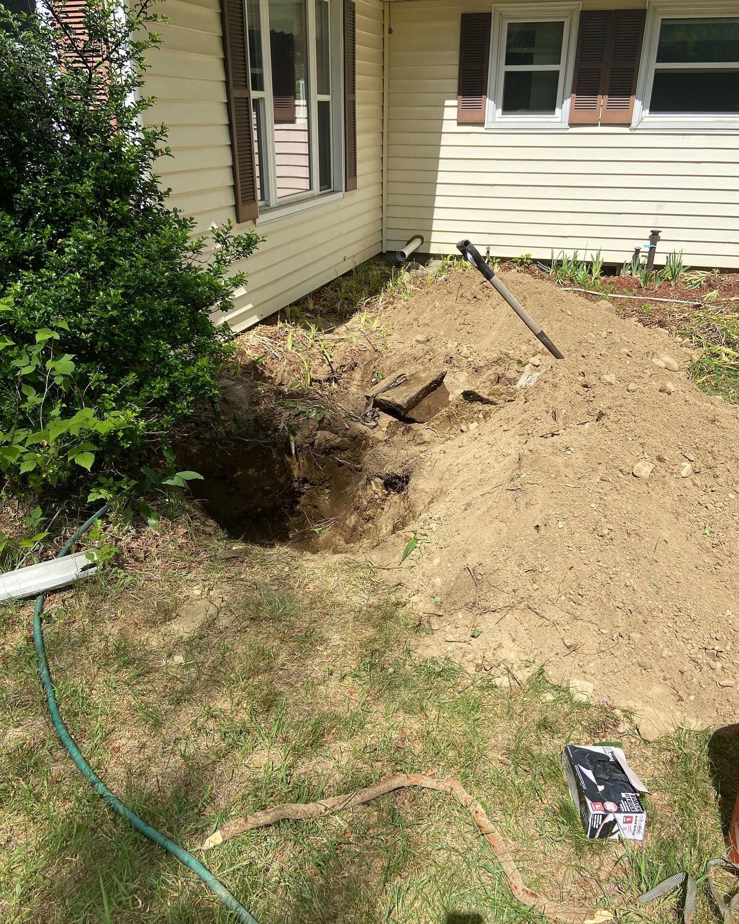A trench has been dug in the soil next to the exterior wall of a house with yellow siding and two windows.