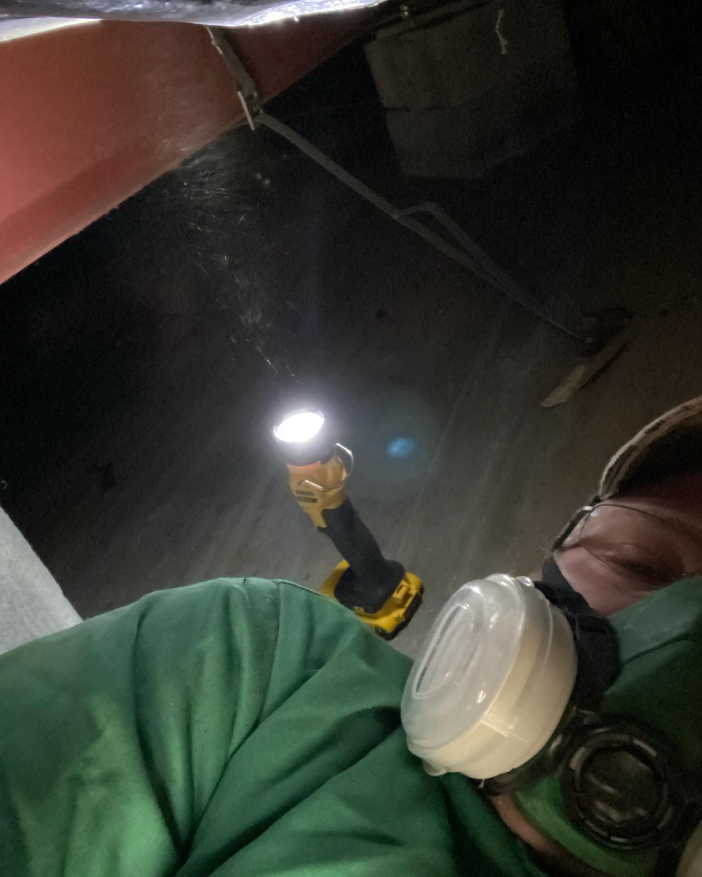 A person wearing a respirator mask works in a dark crawl space, illuminated by a yellow battery-powered LED light.