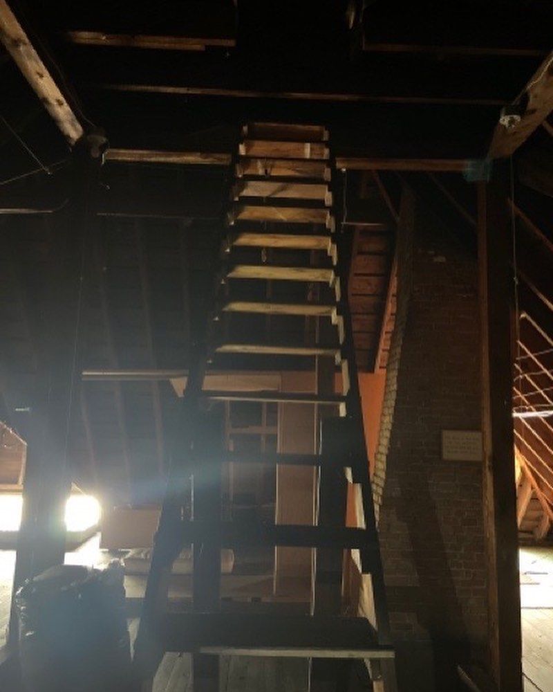 A wooden ladder stands in a dimly lit attic, leading up to an opening in the dark rafters.