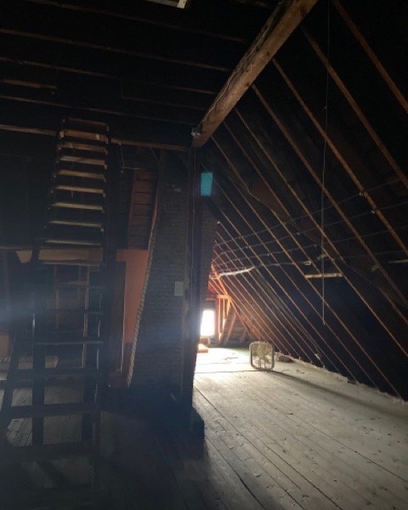 A dim attic space with wooden flooring, slanted rafters, a central chimney, a wooden ladder, and a fan near a light source.