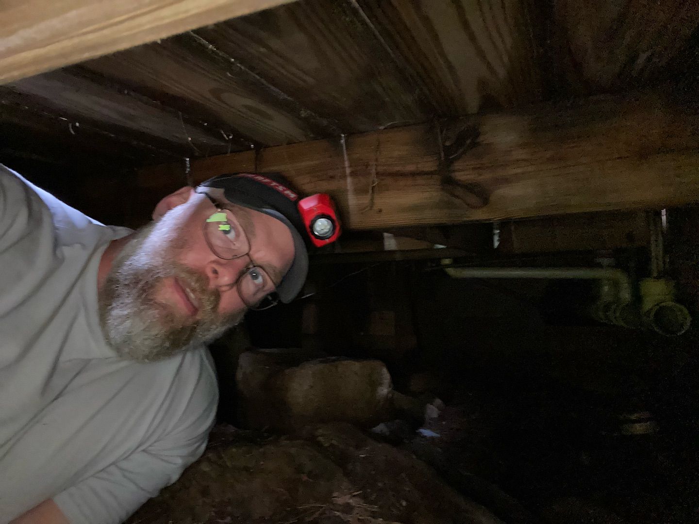 A bearded person wearing a headlamp looks at the camera while lying in a dark crawl space under wooden floor joists.