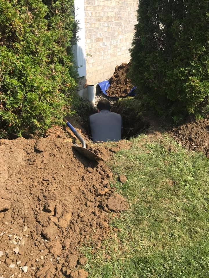 A person working in a hole dug into the ground next to a house foundation and green shrubs, with a shovel nearby.