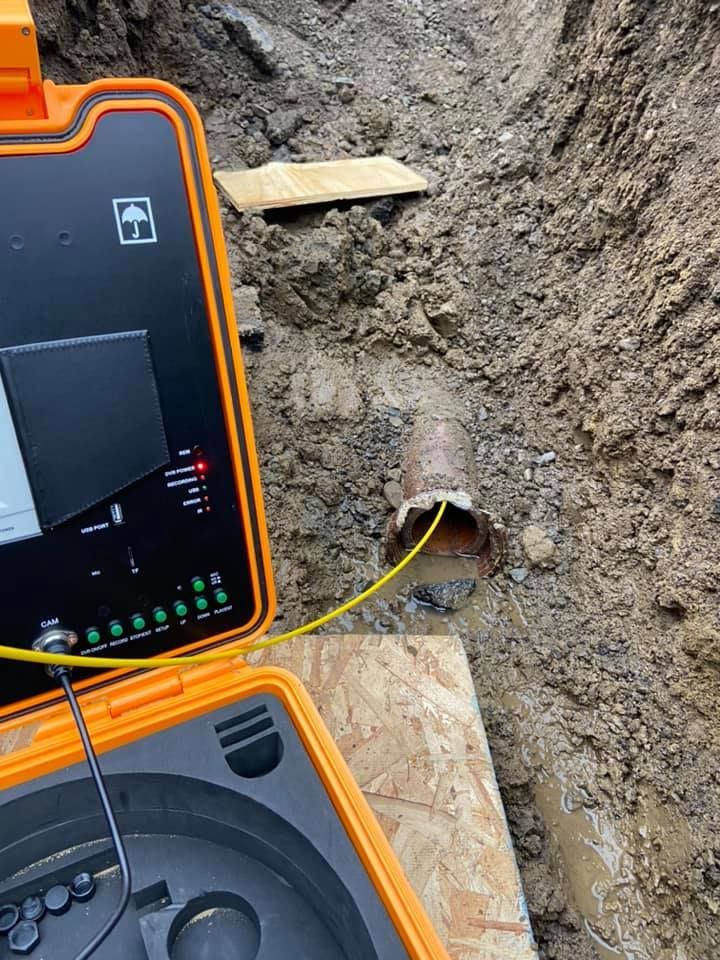 A yellow pipe inspection camera unit sits on a wooden board with its cable inserted into an exposed pipe in a trench.