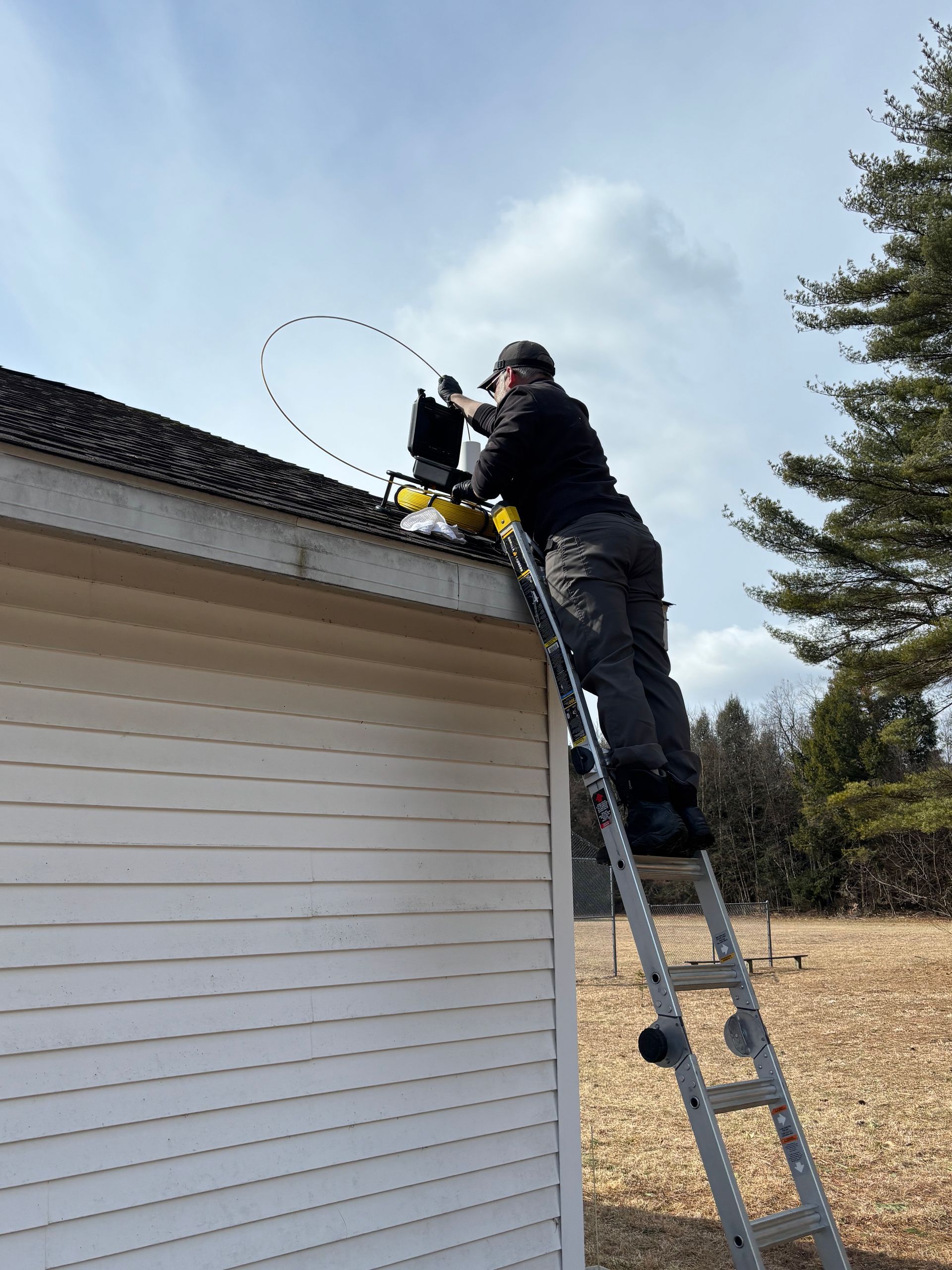 A person standing on an extension ladder works on a roofline while feeding a long, curved cable into an opening.