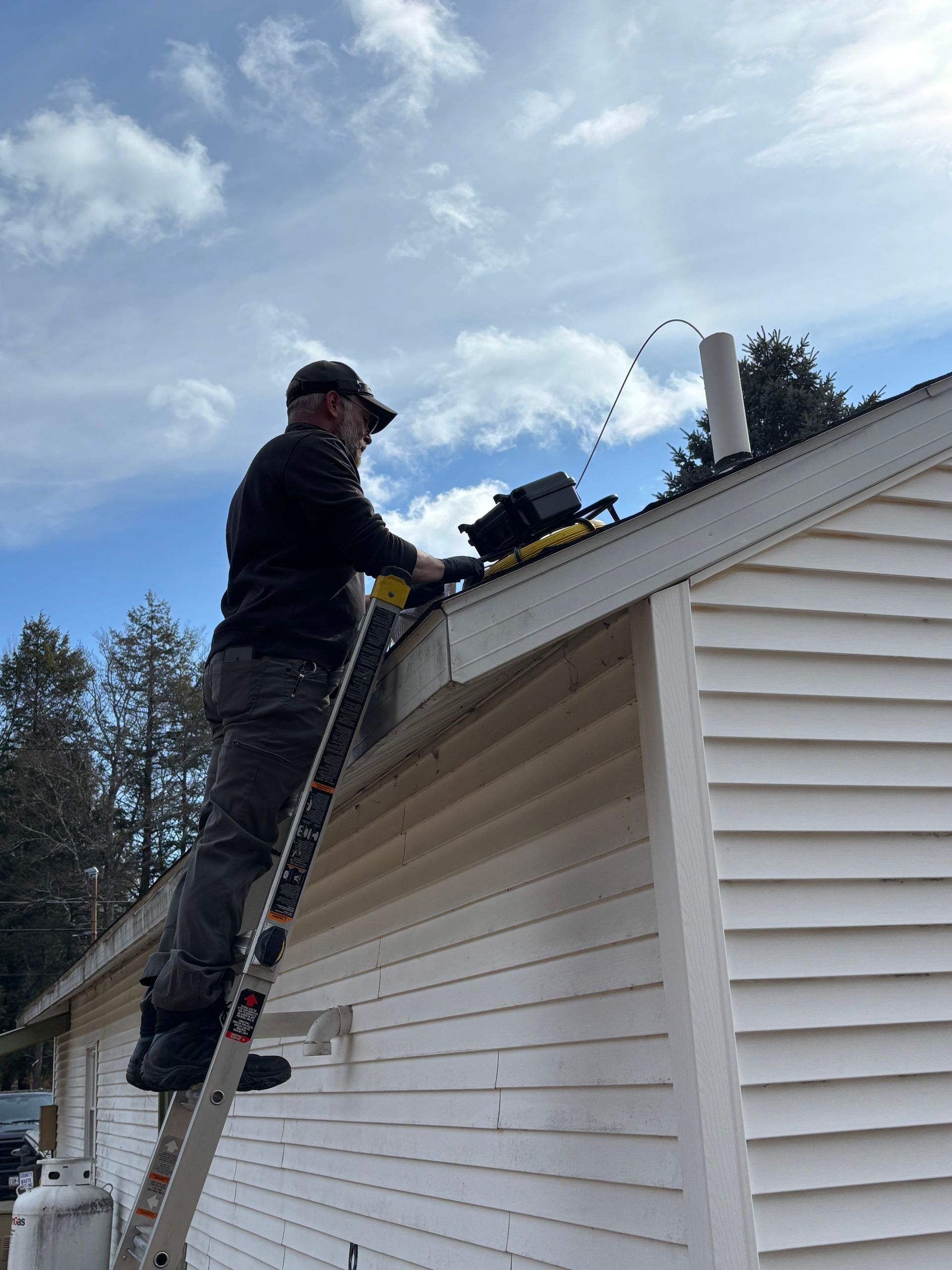 A person wearing a dark jacket and baseball cap stands on an extension ladder while working on a house roof.