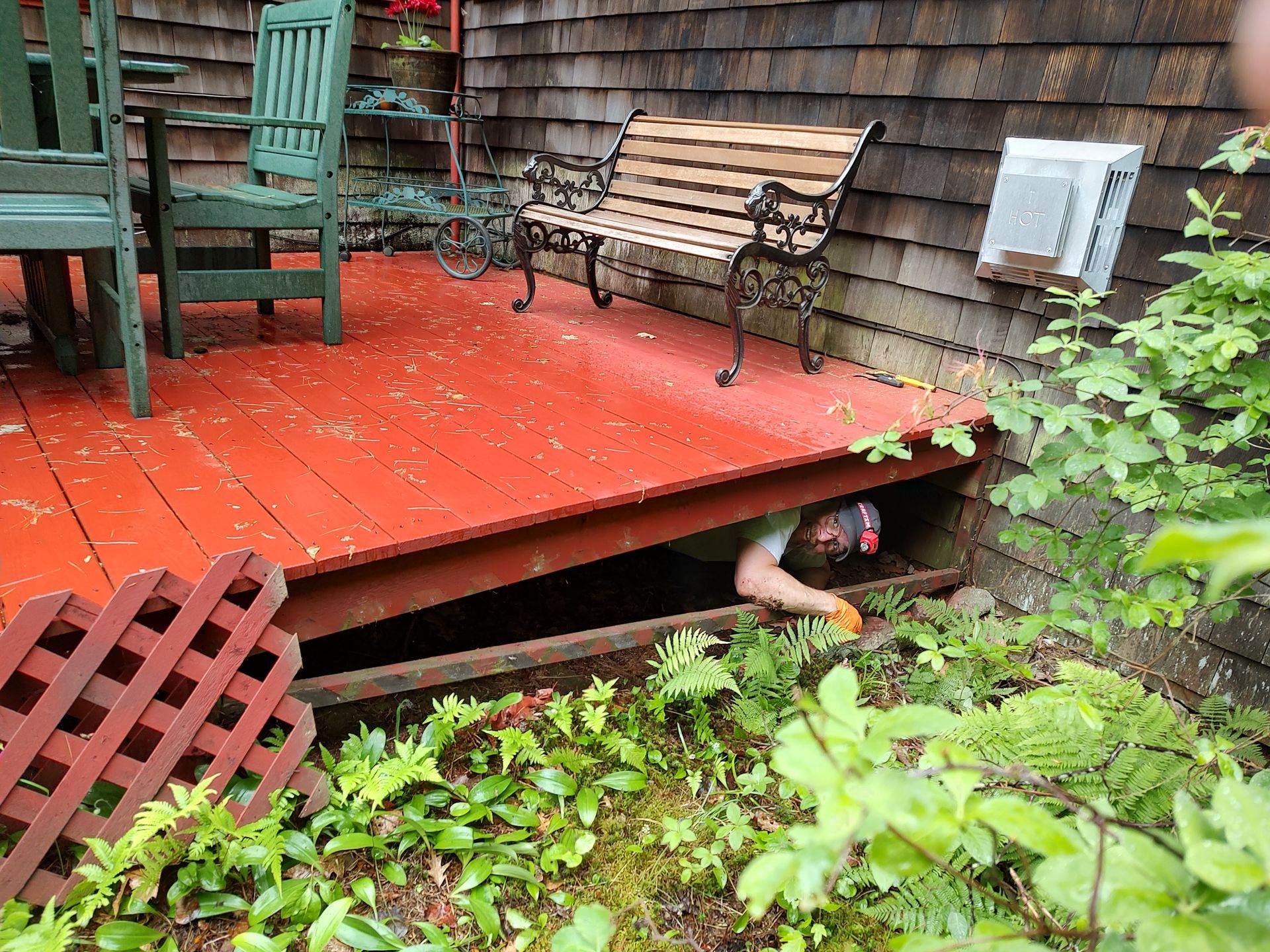 A person wearing gloves reaches into the dark crawl space under a red wooden deck, near a bench and outdoor furniture.