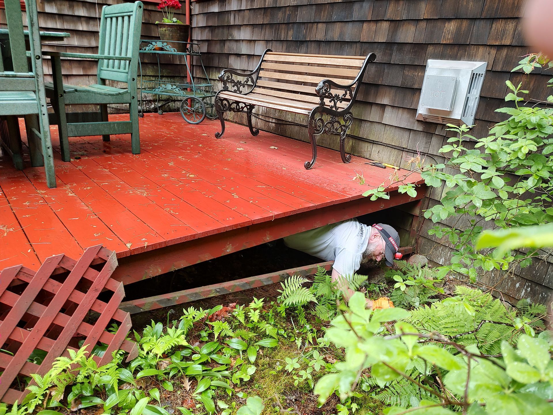A person in a gray shirt crawls into the dark space beneath a painted red wooden deck in a yard.