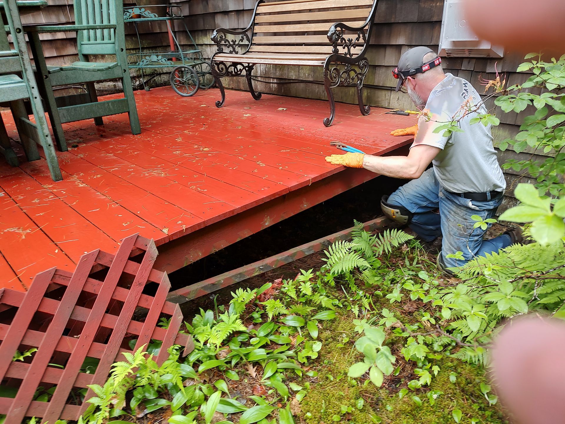 A person wearing a headlamp and gloves repairs the edge of a red wooden deck, with a bench and chairs nearby.