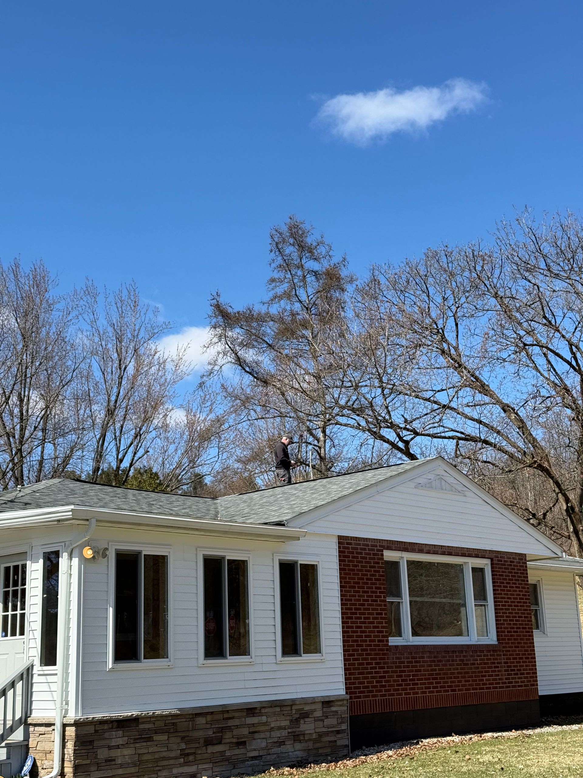 A white, single-story house with a brick facade and gray shingles, surrounded by bare trees under a bright blue sky.