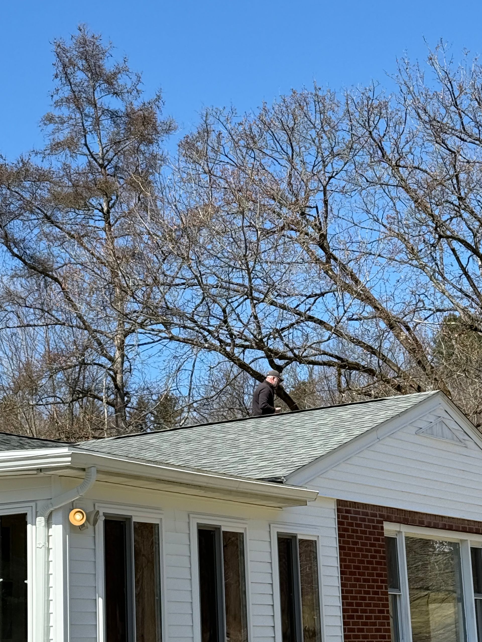 A person working on the shingled roof of a white house on a sunny day with bare trees in the background.