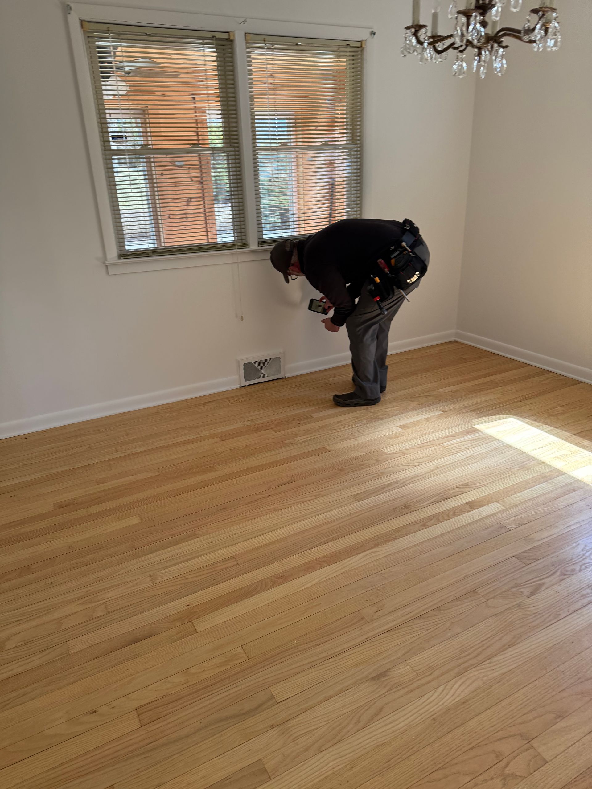 A worker in dark clothing inspects a wall near a floor vent in a room with hardwood floors and wooden blinds.