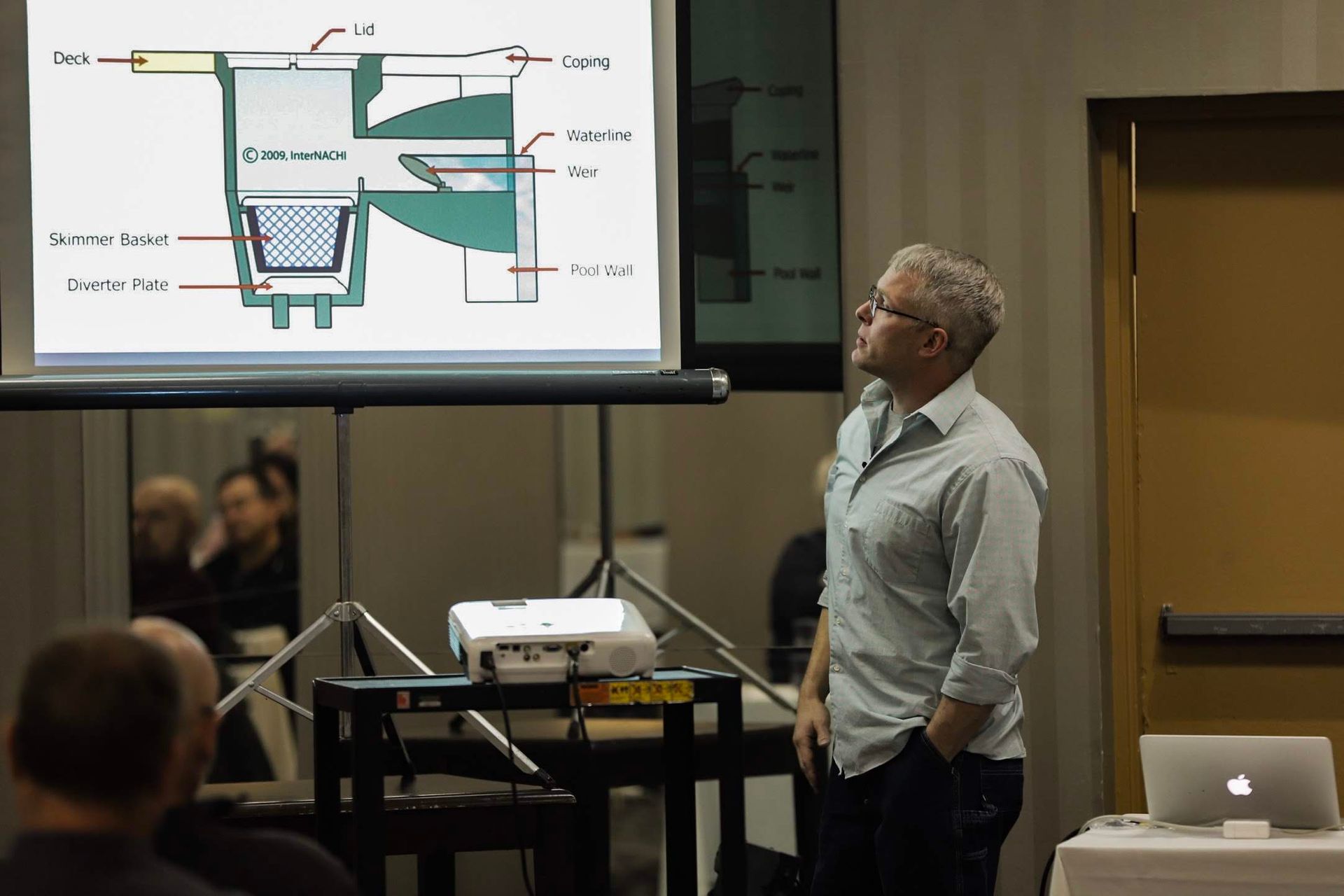 A presenter in a light button-down shirt stands before a screen displaying a technical diagram of a machinery component.