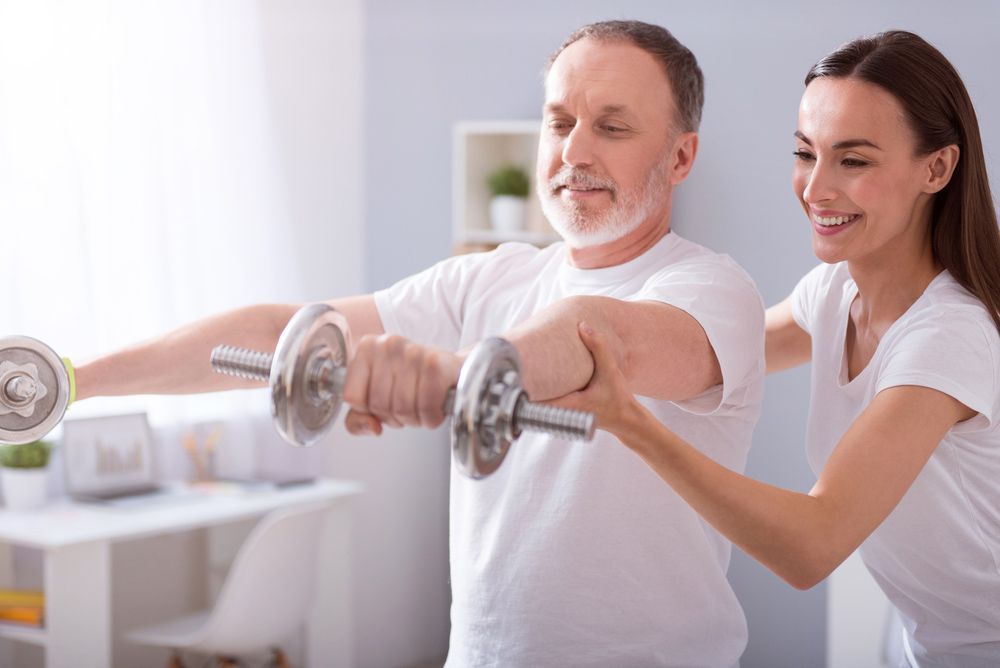 A Woman Is Helping An Older Man Lift Dumbbells — Caves Coastal Physiotherapy in Swansea, NSW