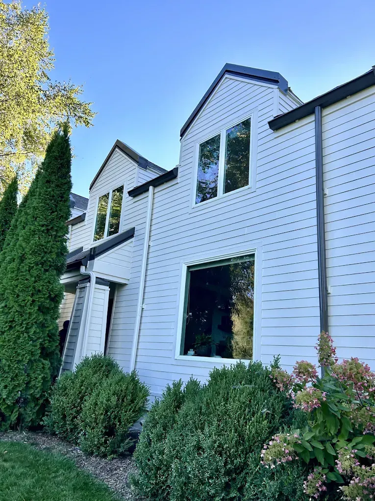 White two-story house with black-trim windows, front shrubs, and tall trees under a blue sky