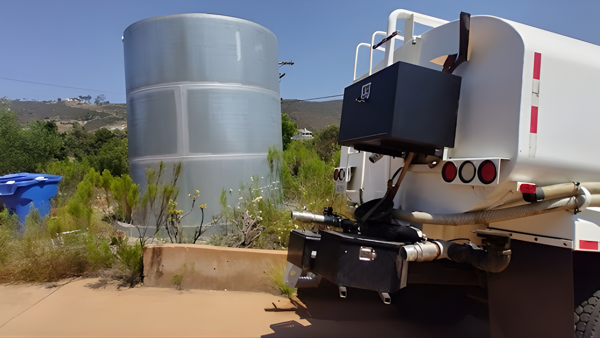 A white water truck parked next to a large, metallic cylindrical storage tank in a sunny outdoor area.