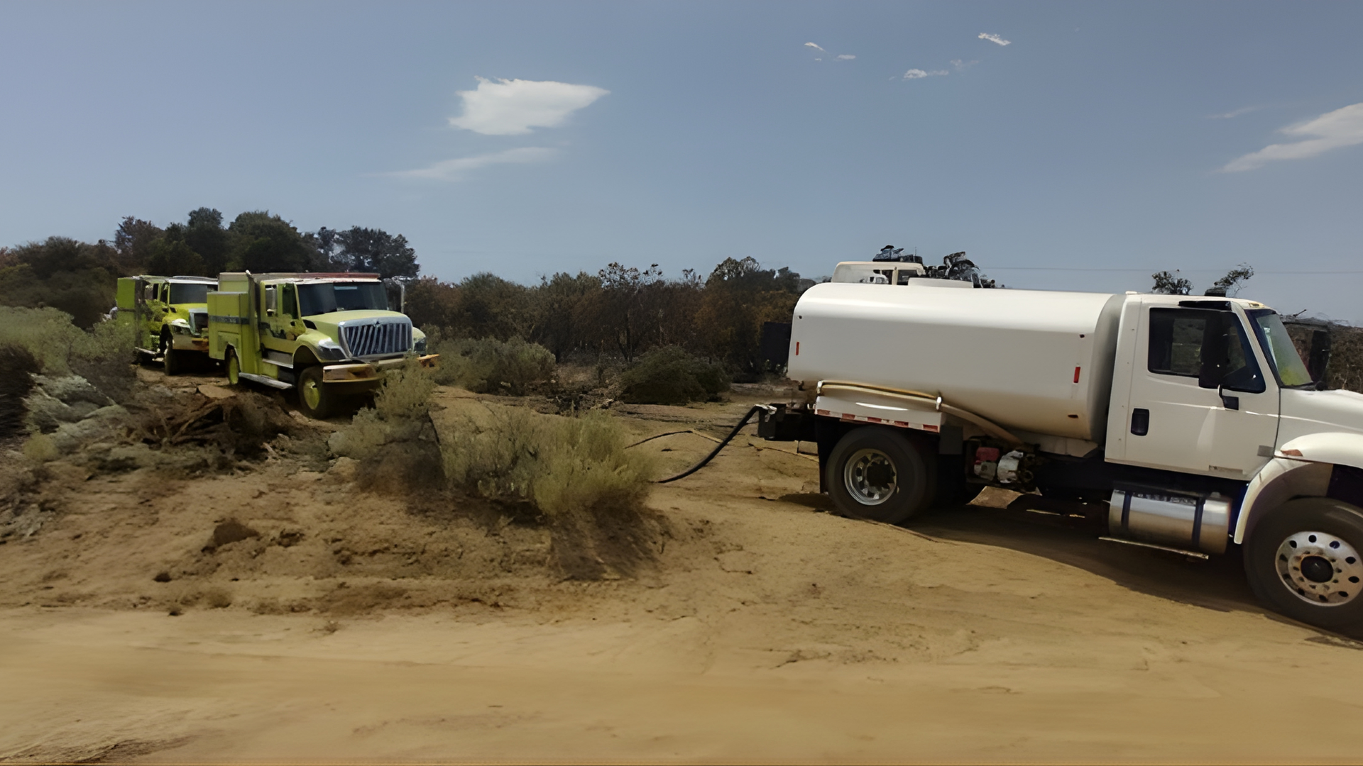 A white water tanker truck connected by a hose to two lime-green fire engines in a dusty, rural field.