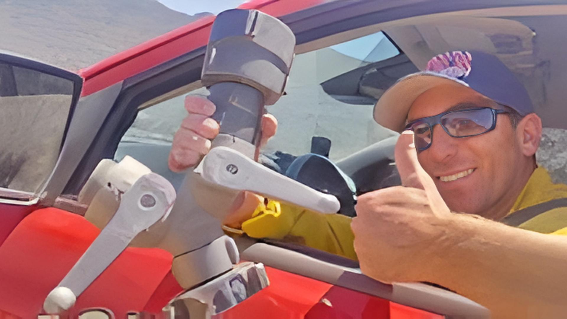 A person in a yellow uniform and cap gives a thumbs up while holding a large fire hose nozzle inside a red vehicle.