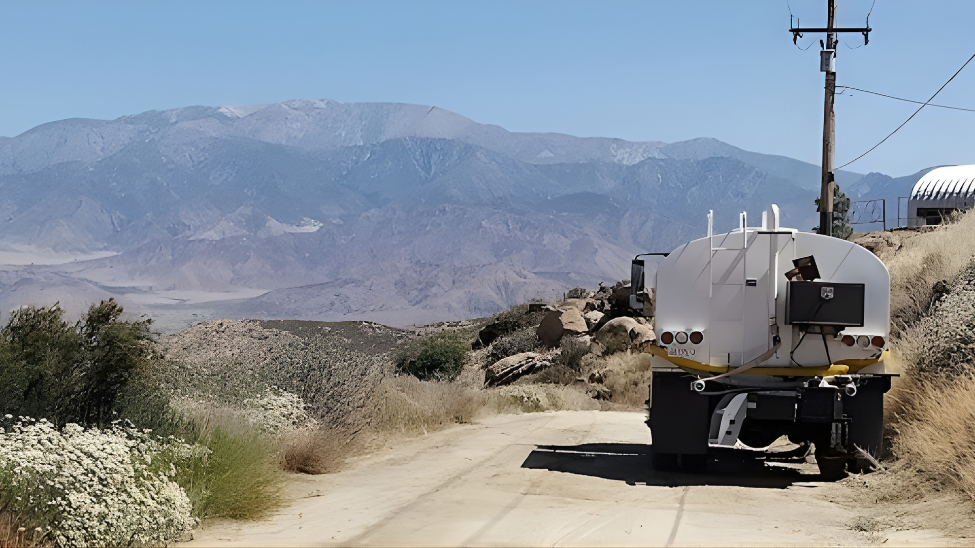 A white water truck parked on a dirt road in a desert landscape with large, arid mountains in the background.