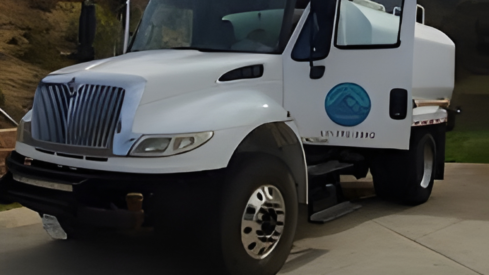 A white International truck with a large water tank and a circular blue logo on the door parked on a paved driveway.