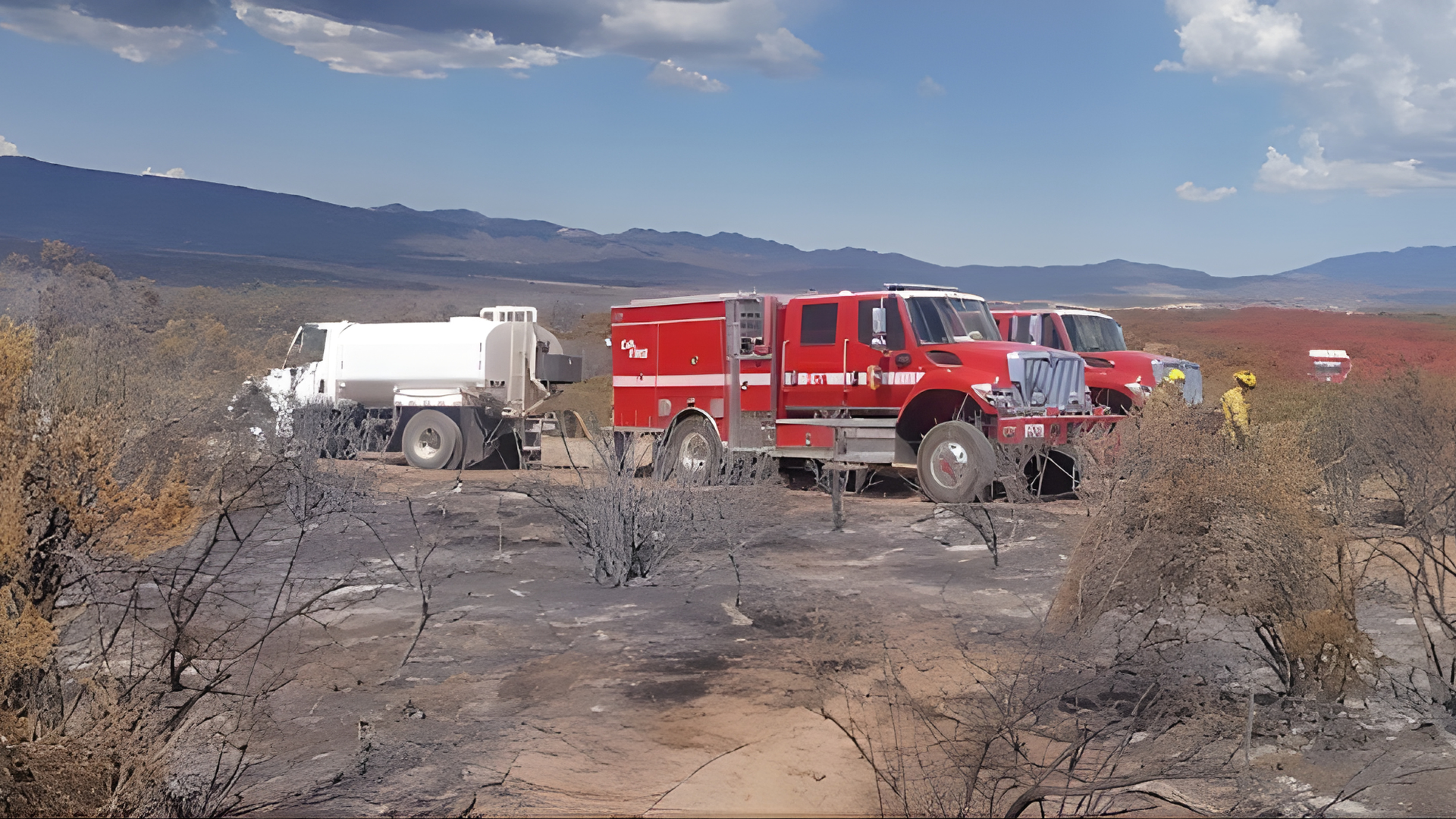 Fire trucks parked on a charred, burnt landscape in a desert area under a partly cloudy sky.