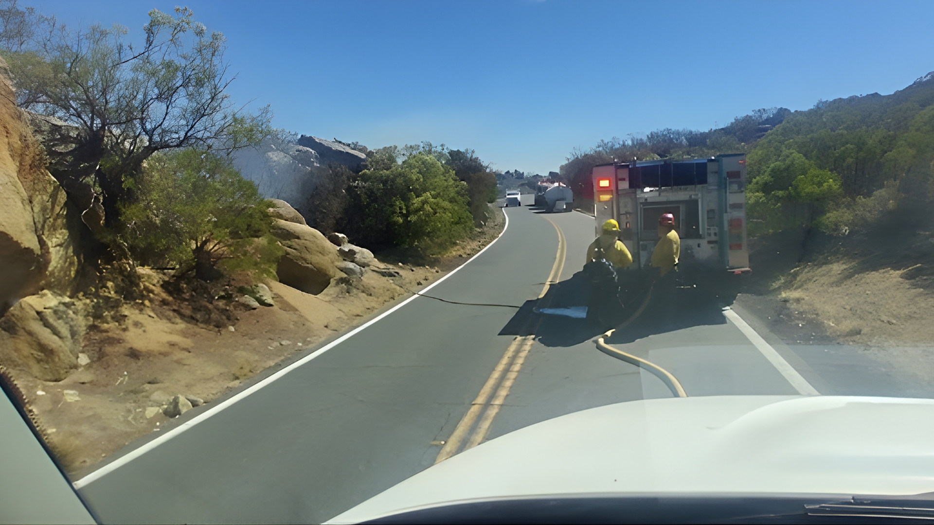 Firefighters in yellow gear stand by a fire engine on a road with smoke rising from the brush on the left.