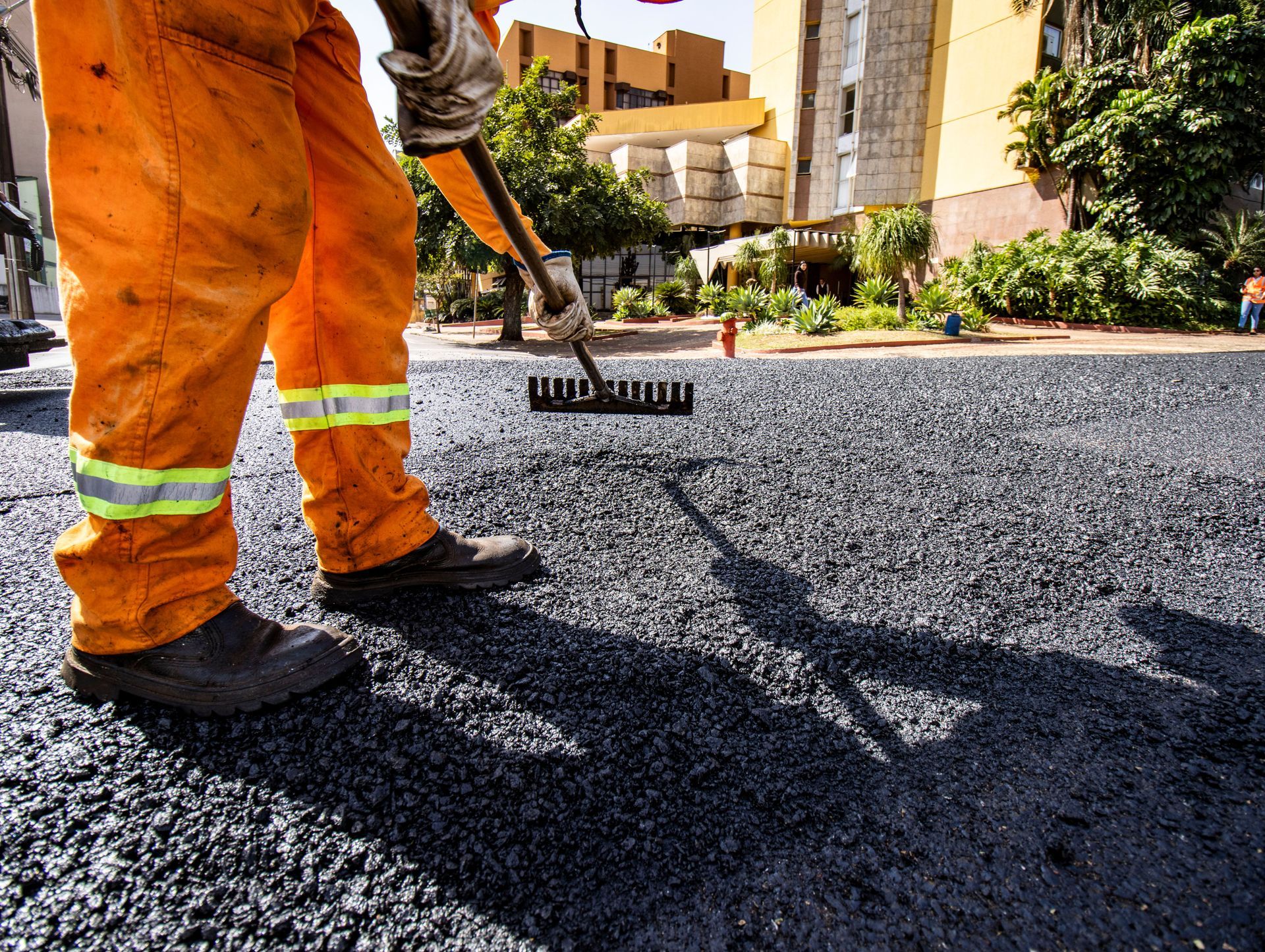 Construction worker smoothing pavement; wearing orange safety vest, white helmet, and boots.